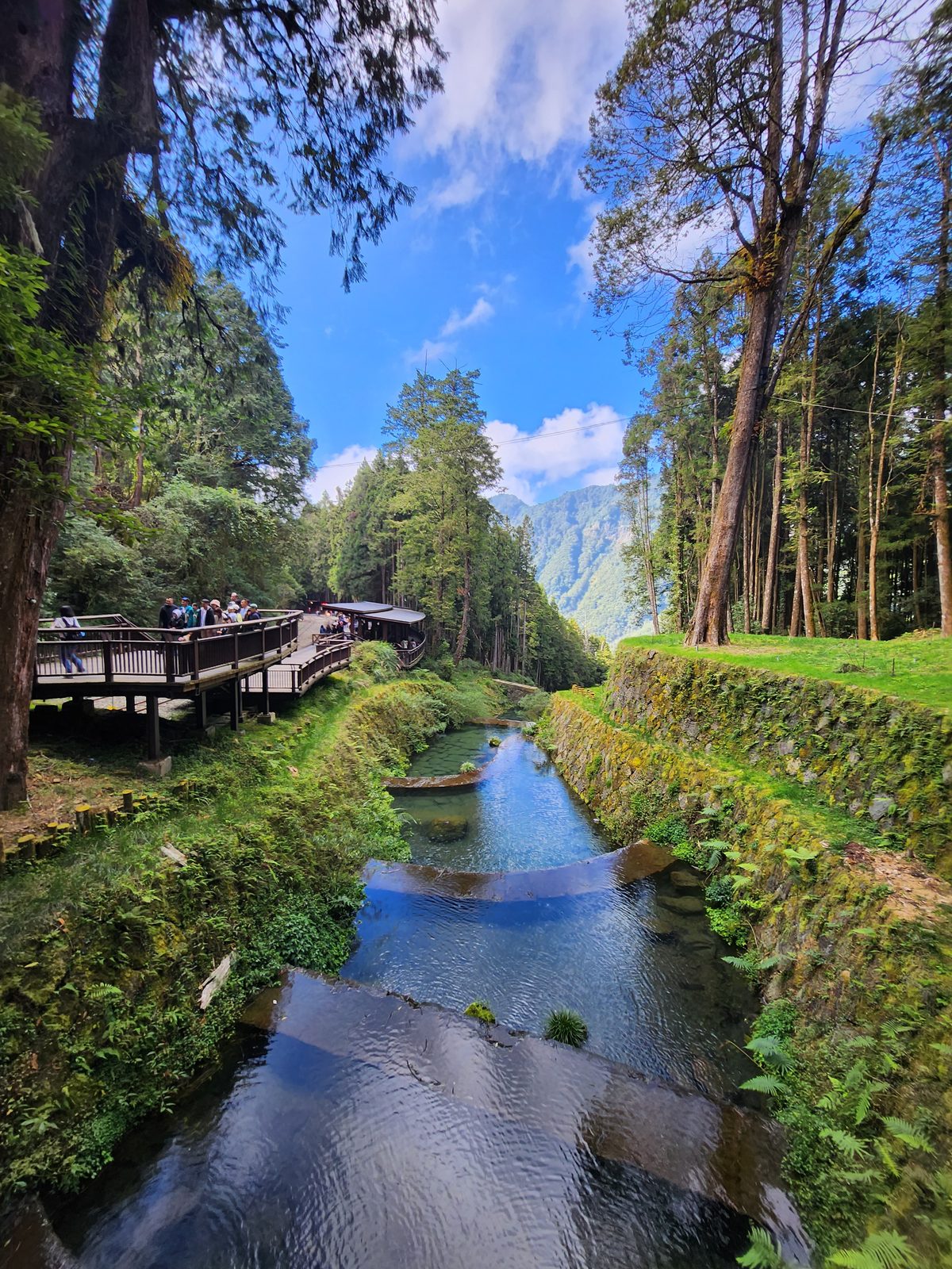 木棧橋橫過水池,遠方是山稜與白雲,棧道上有零星旅客