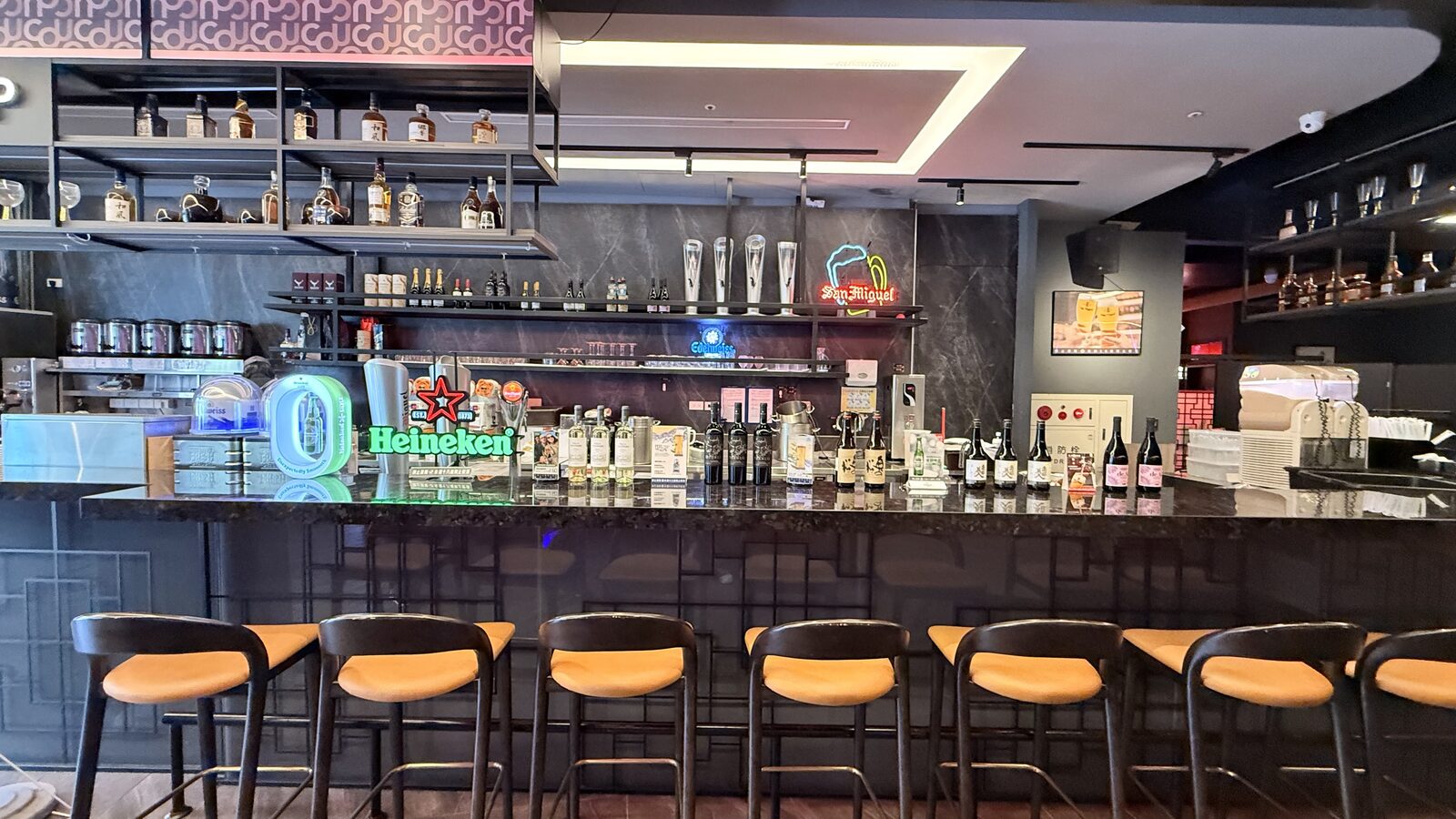 술장, 맥주 네온사인, 음료 제조 설비가 있는 긴 바와 앞에 줄지어 놓인 주황색 쿠션 하이 체어