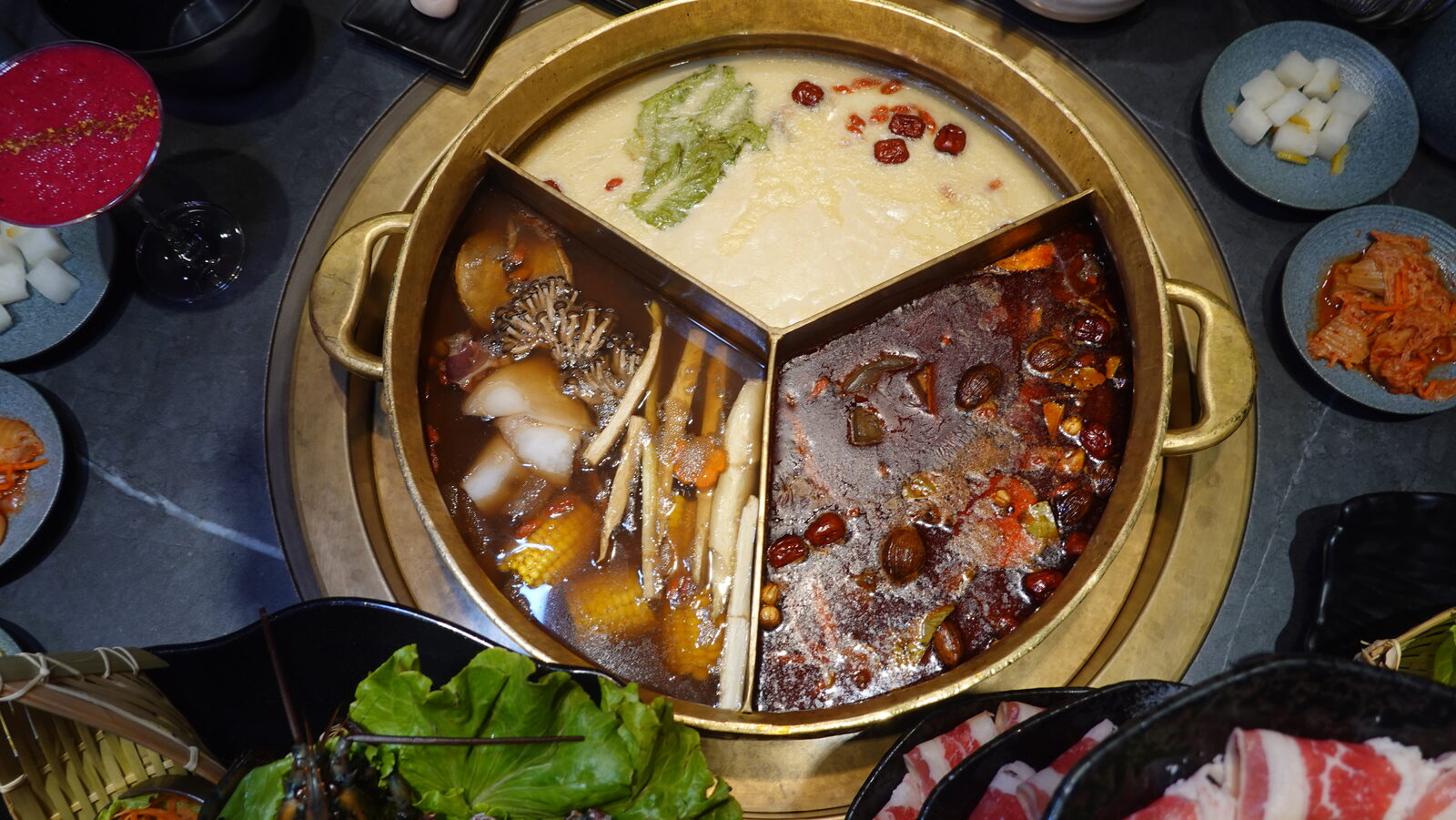 붉은 마라 육수, 뽀얀 백탕, 맑은 육수가 나란히 담긴 COUCOU 3구 황동 냄비를 위에서 본 모습과 주변의 반찬 접시