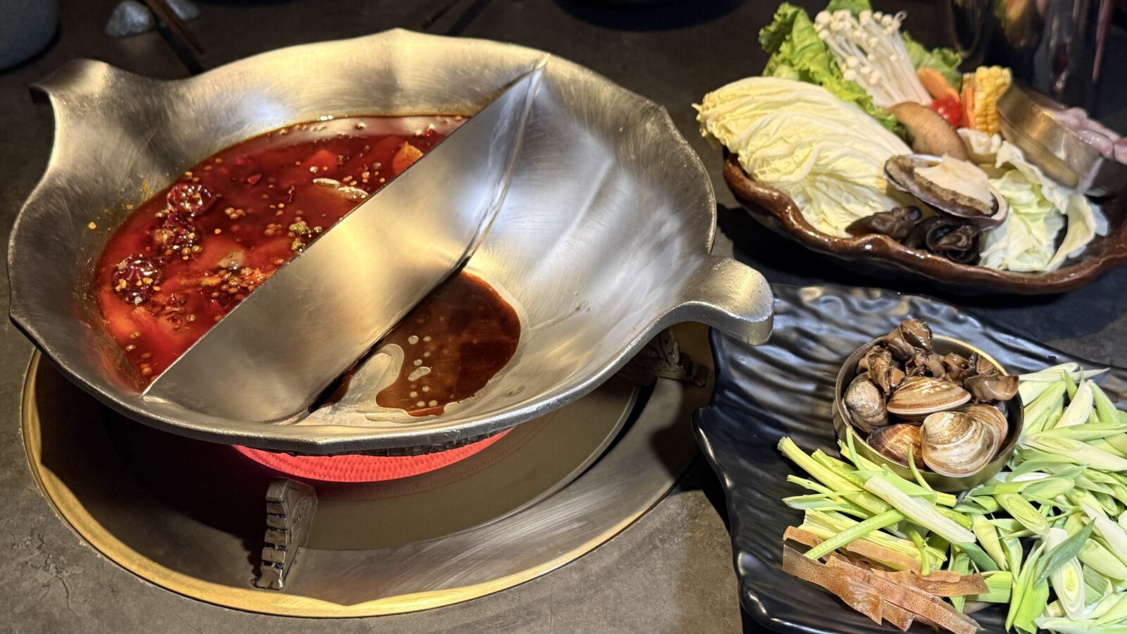 한쪽은 붉은 마라 육수, 빈 다른 쪽에는 짙은 색 소스를 먼저 부은 2구 냄비와 옆 검은 접시의 마늘대·마른오징어 채, 작은 구리 그릇의 소라살과 바지락