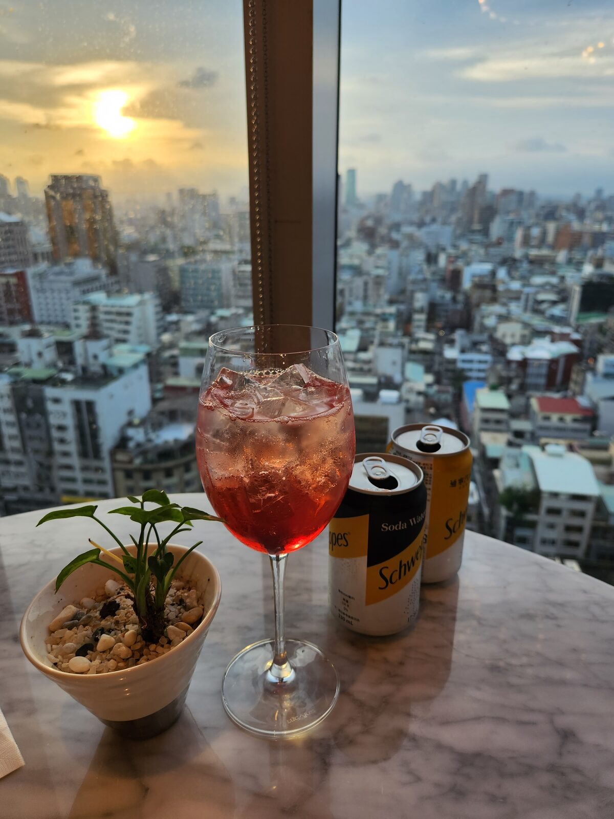 高腳粉紅飲品與 Schweppes 蘇打水,背景落地窗日落市景