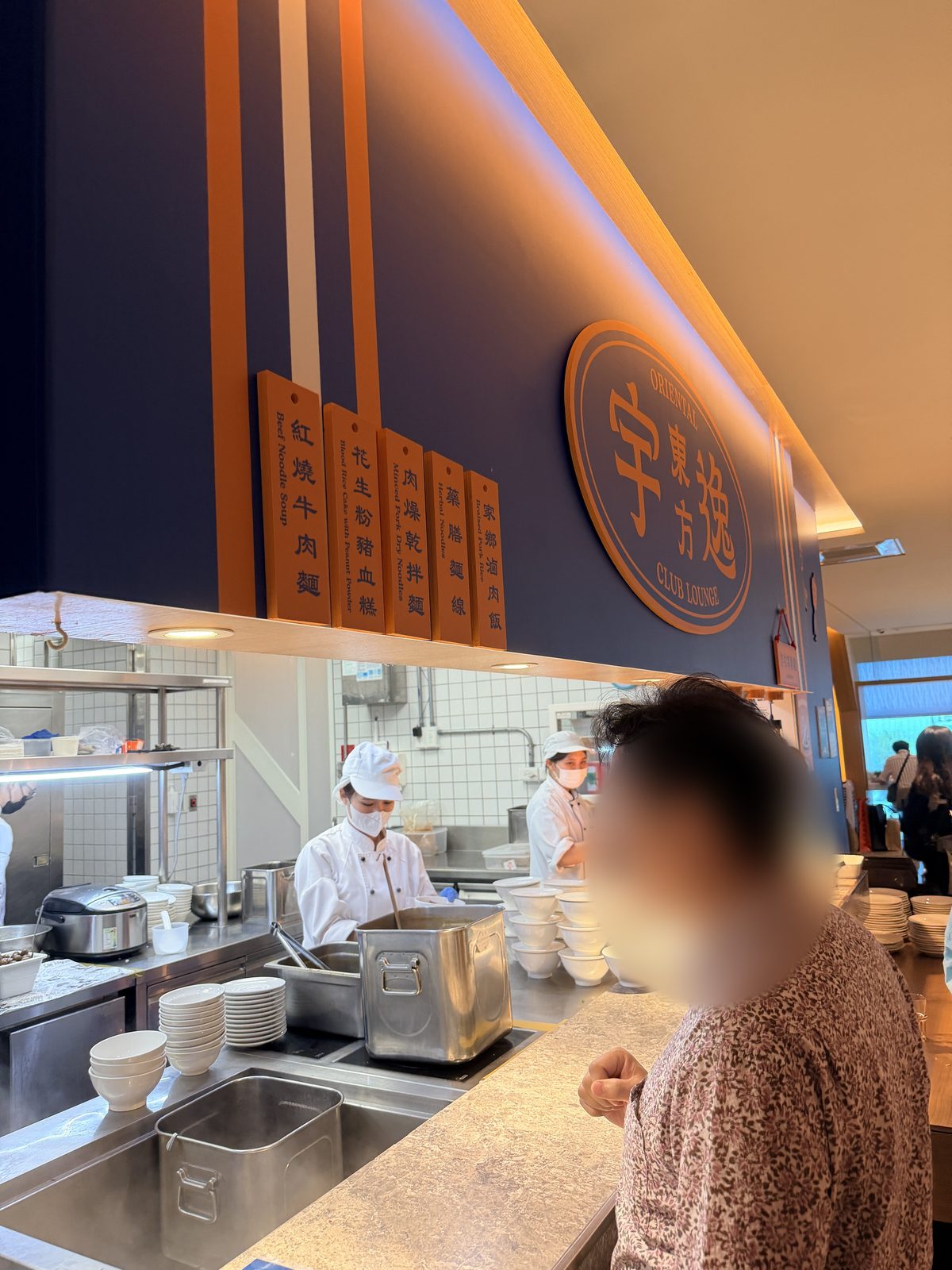 木製の品名札が上に掛かる注文カウンターと、奥で調理するスタッフ。手前に利用者が一人