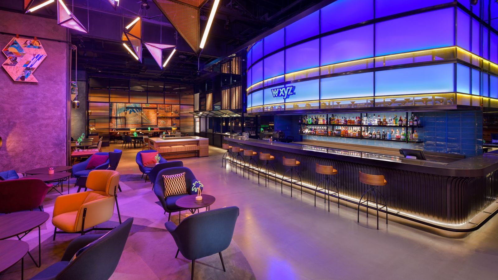 A long bar counter and high stools under purple-blue lighting, with several colorful armchairs in front