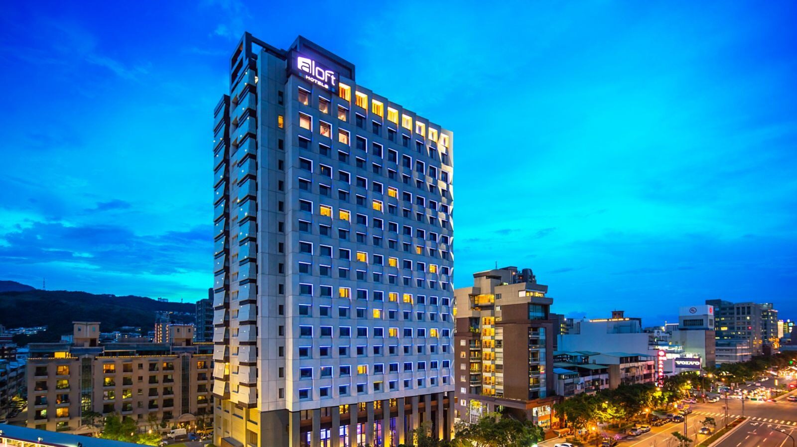 The Aloft tower lit up at dusk, with blue sky and distant mountains behind