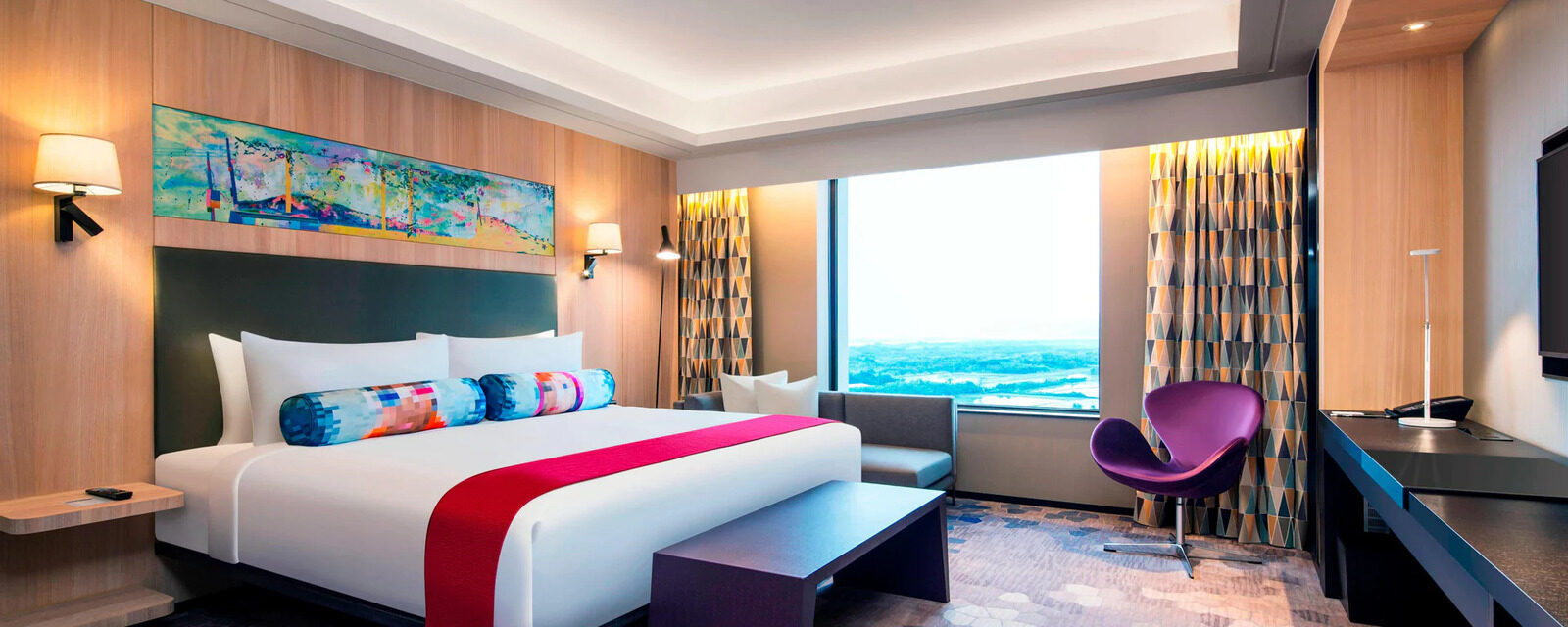 A king bed with a red bed runner in a suite bedroom, with a purple armchair and desk by the window