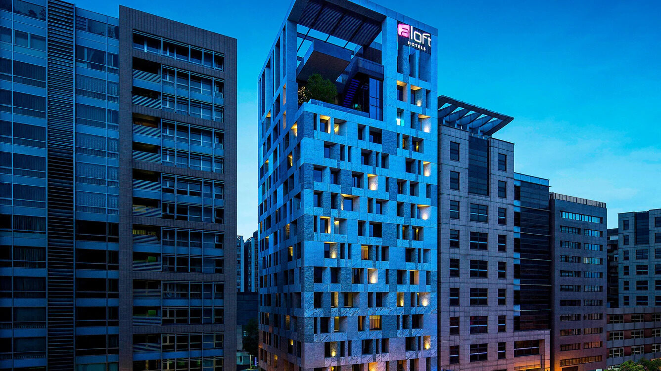 The Aloft building lit up at dusk with the aloft sign on the roof, flanked by office towers