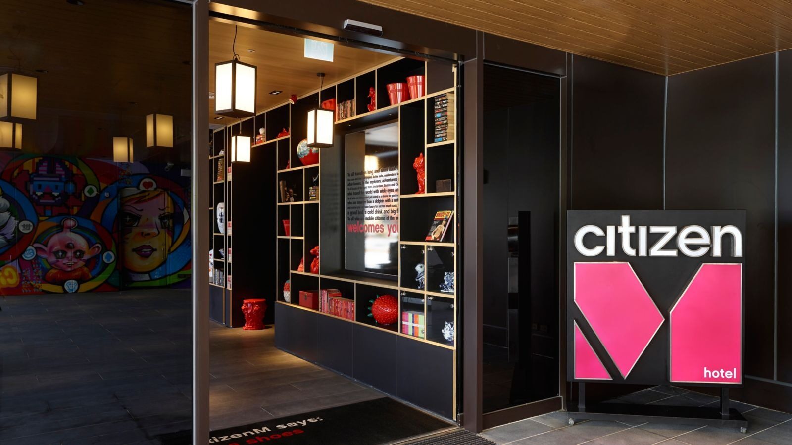 A citizenM sign with a pink M beside the entrance, and red-and-black bookcases full of art objects inside the door