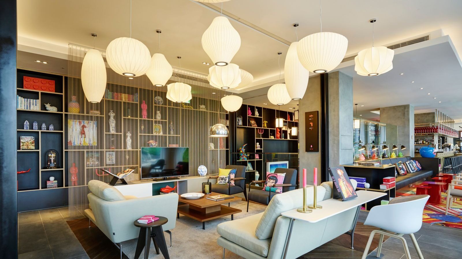White sofas and coffee tables in a bright communal living room under white globe lights, with bookcases of art objects along the wall