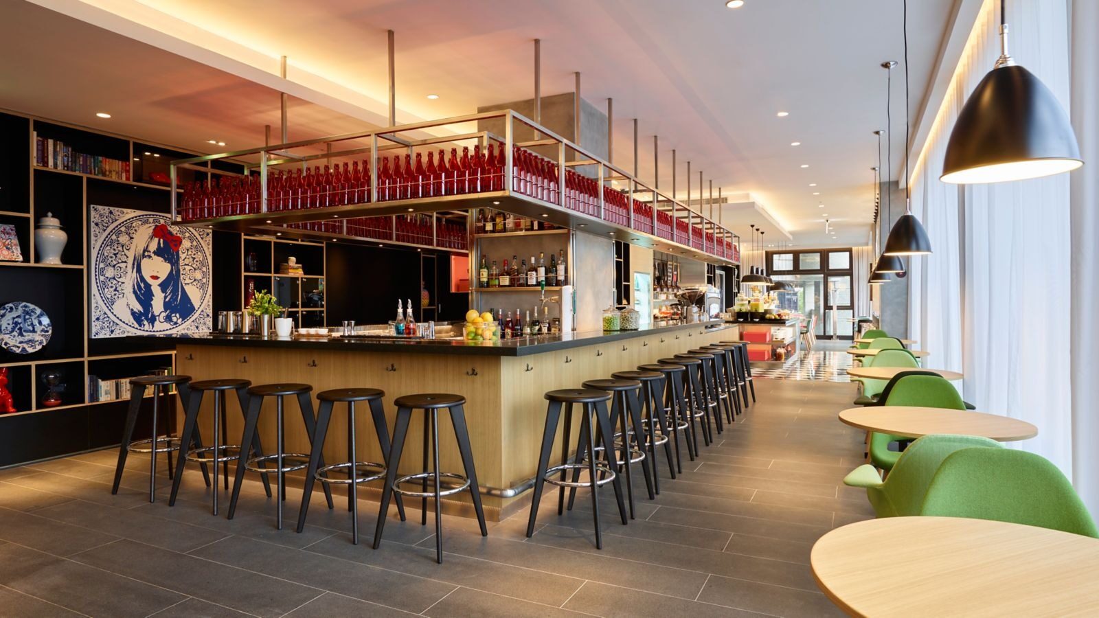A row of bar stools at a long counter, shelves of red bottles and jars above it, with green chairs and round tables alongside