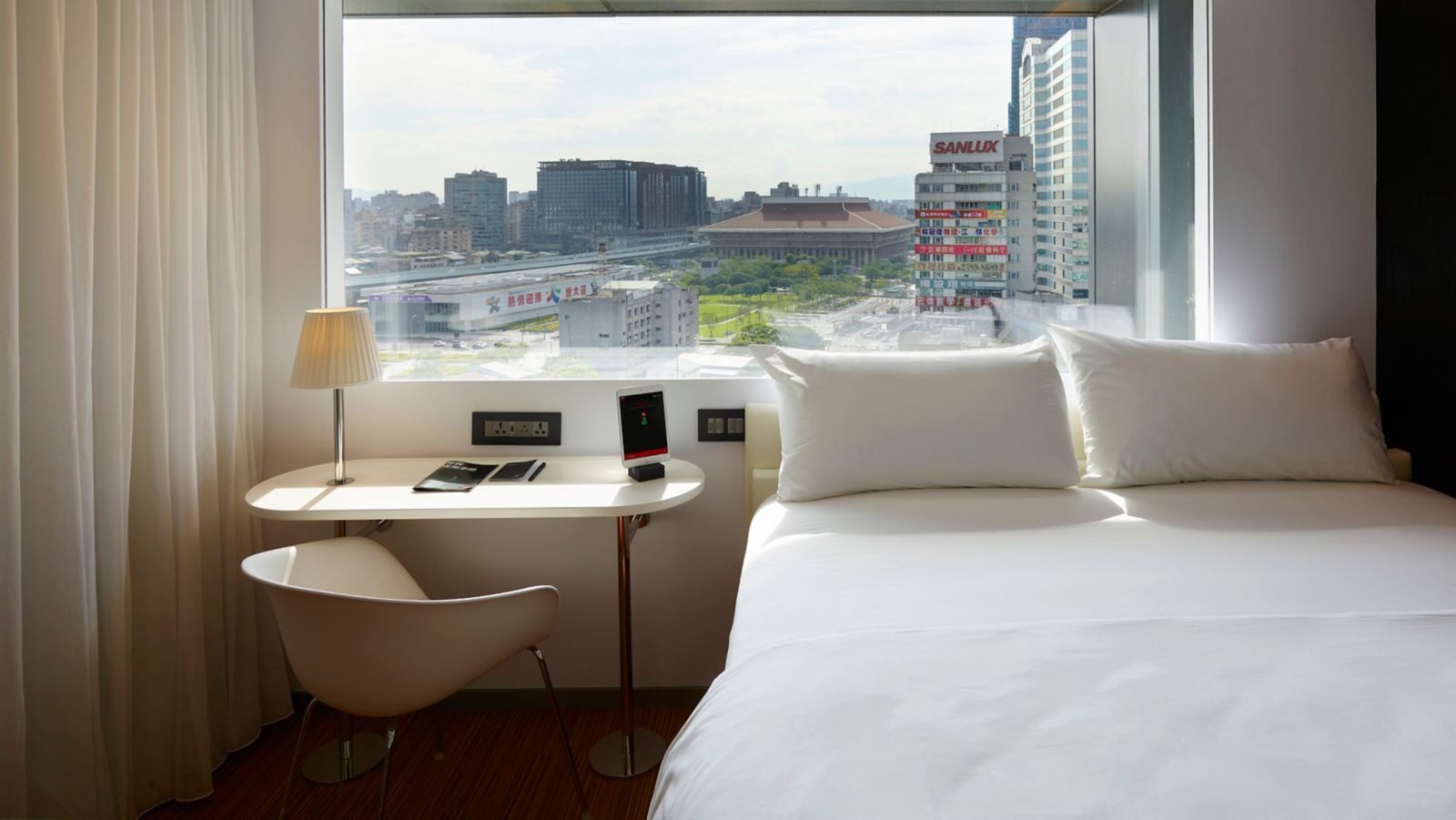 A desk and lamp beside a large bed, facing a wall-wide window