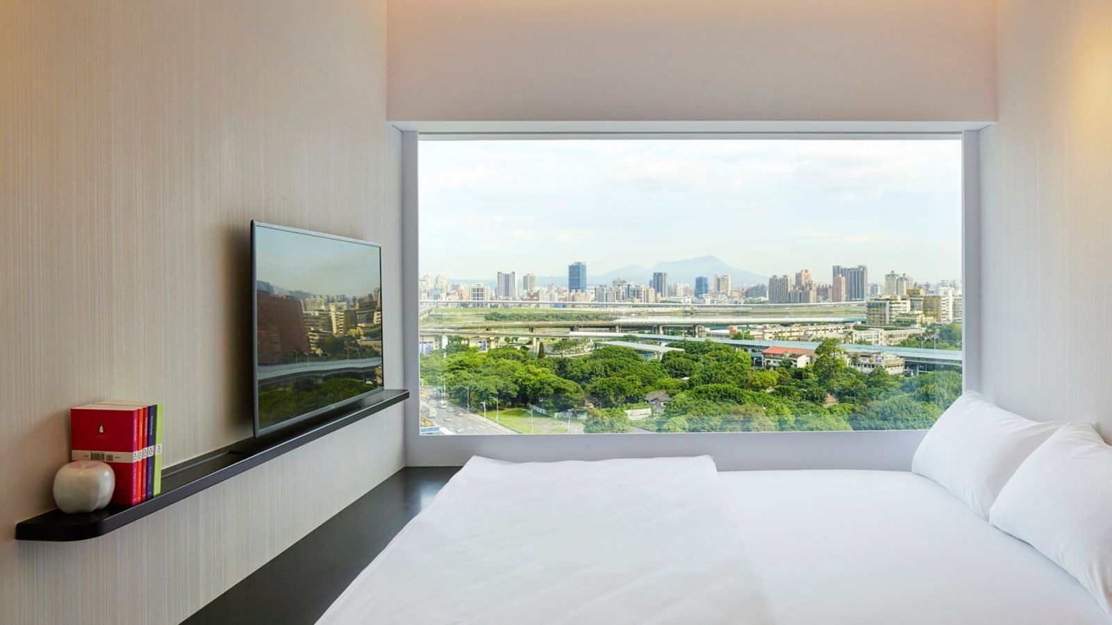 White bedding facing a wall-wide window, with nearby trees, an elevated road and the distant city and mountains outside