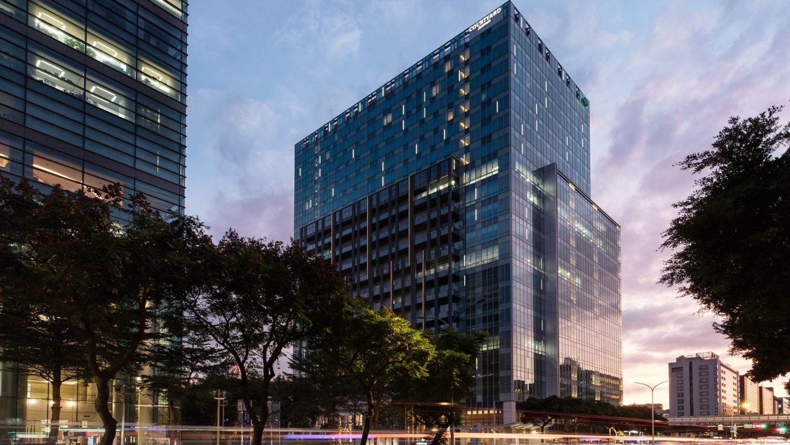 The glass tower at dusk, with street trees and streetlights in the foreground