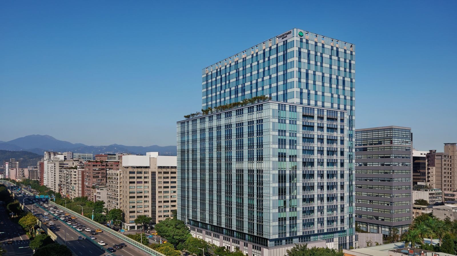 A glass tower under blue sky beside a busy boulevard, with mountain ridges in the distance