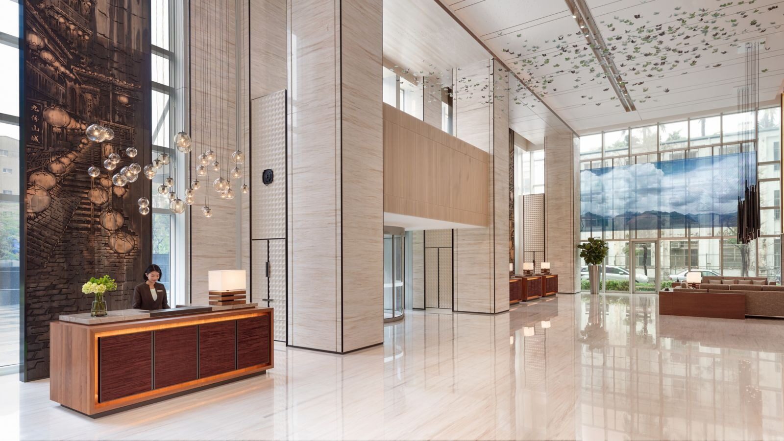 A wooden reception desk in the high-ceilinged lobby, beside a large relief wall of Jiufen's lantern-lit hillside streets and floor-to-ceiling windows