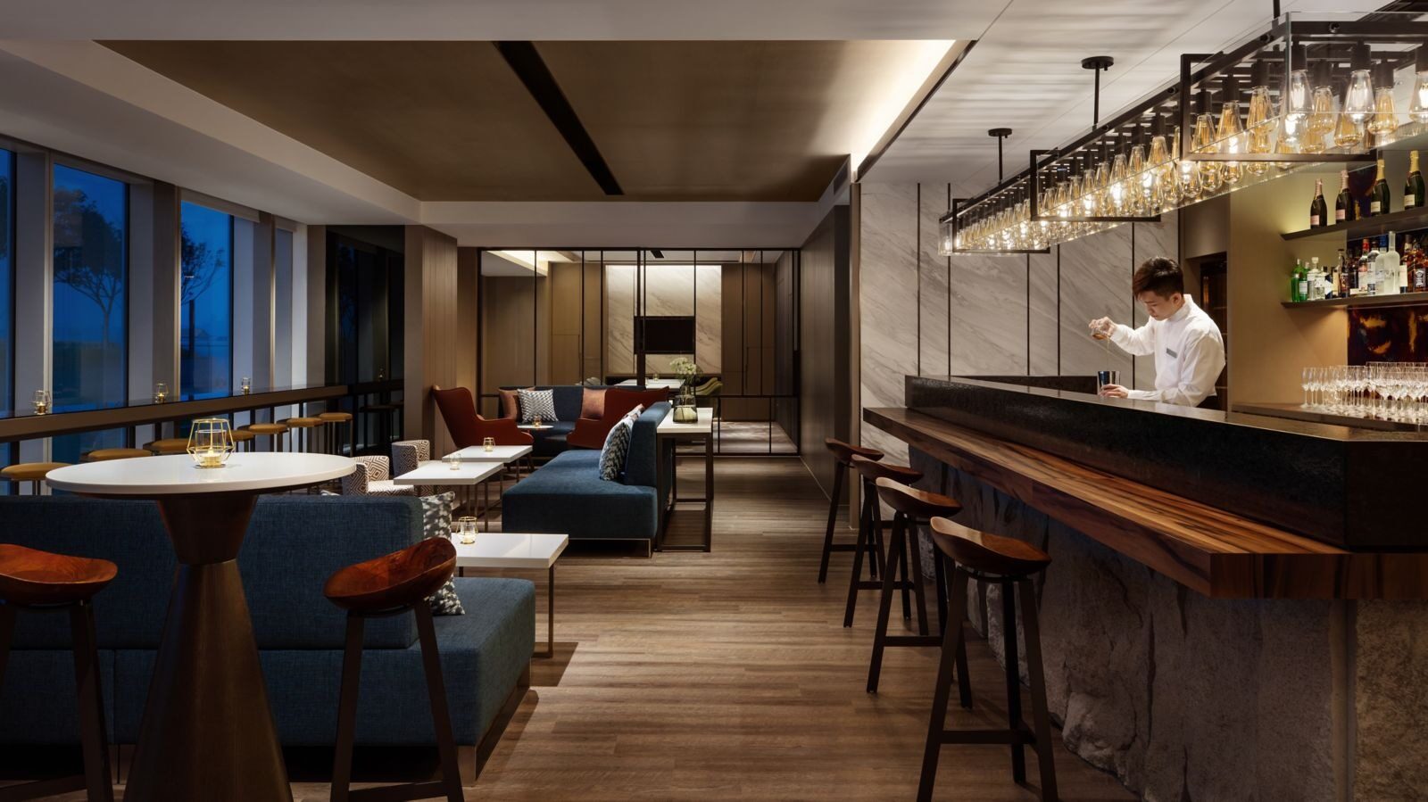 A bartender mixing drinks behind the bar under a row of bulb pendant lights, with blue sofa seating and window views alongside