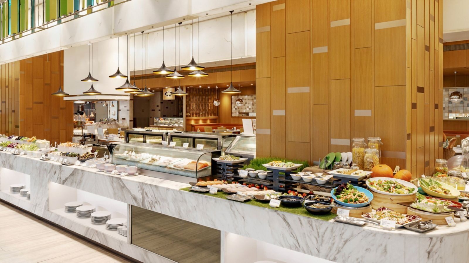 Rows of cold cuts, salads and hot dishes on a white marble buffet counter, with wood paneling and pendant lights behind