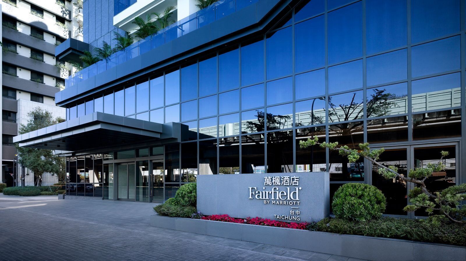 酒店入口的玻璃帷幕与雨遮，前方石墙上刻着 Fairfield 字样，周围种着绿植