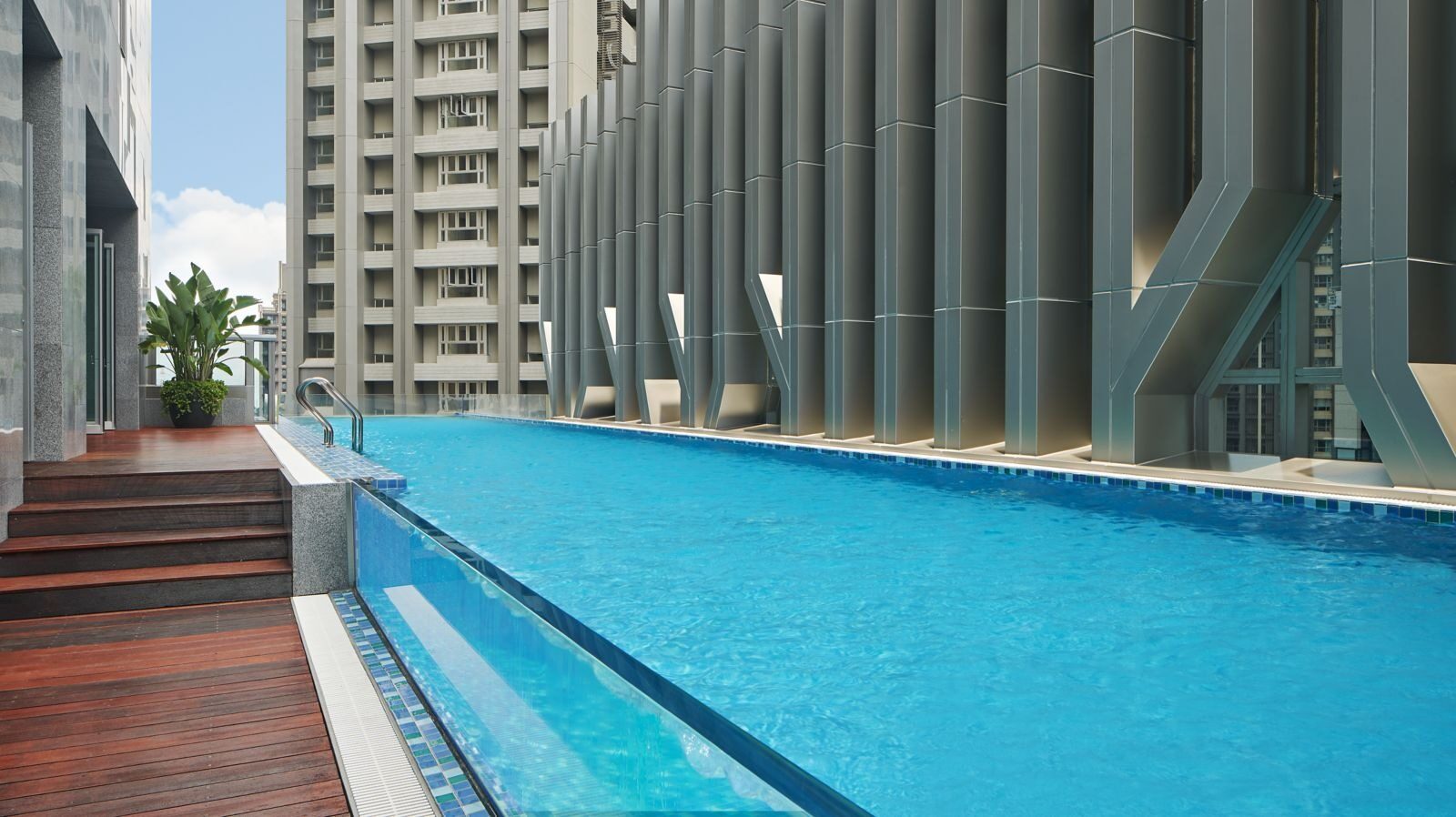 A long infinity pool beside a tower, with a wooden deck and green plants at the edge
