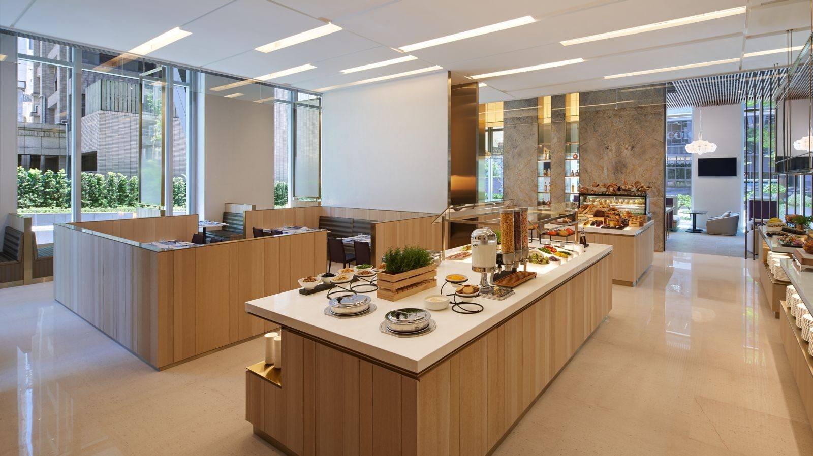 A wooden buffet counter in the center of a bright restaurant, set with bread, cold cuts and green plants