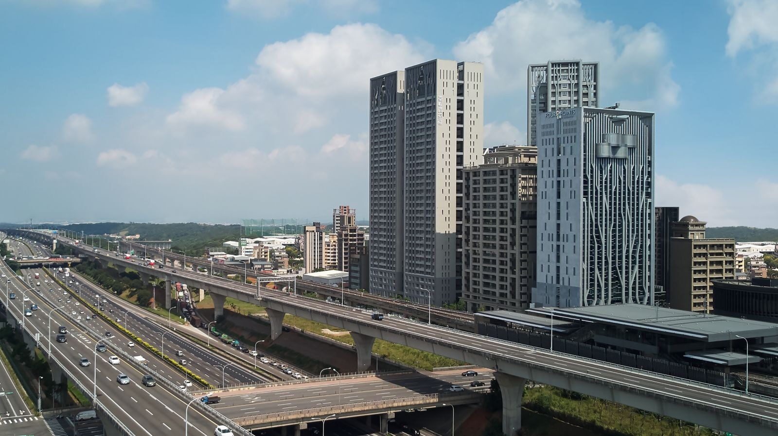 A cluster of towers under blue sky beside a multi-lane freeway and an elevated rail line