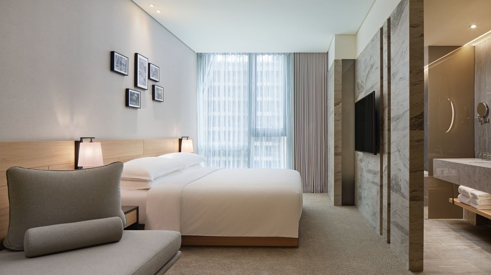 White bedding and a gray bedside sofa in a king room, with a stone feature wall and TV on the right and the bathroom sink behind