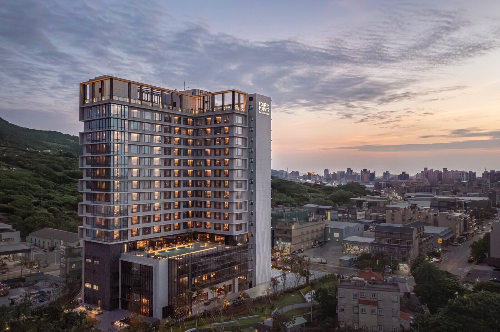 An elevated view of the hotel tower at dusk, surrounded by low-rise streets with city lights in the distance