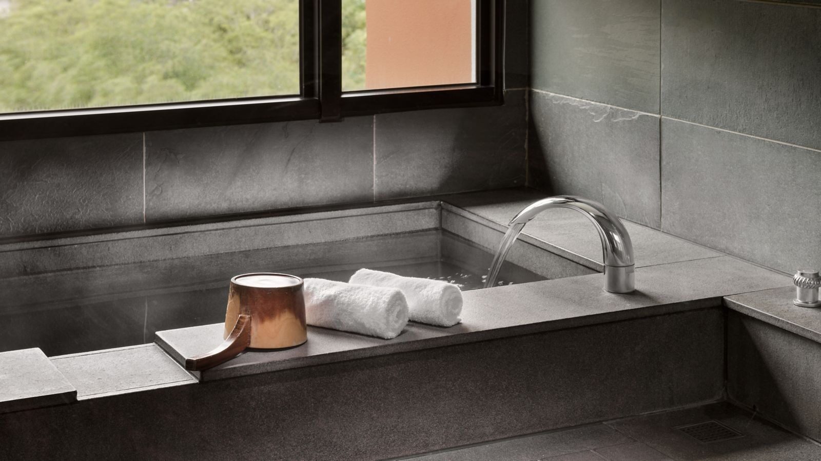 A square gray-stone bath with a towel and copper ladle at the edge, green trees beyond the window