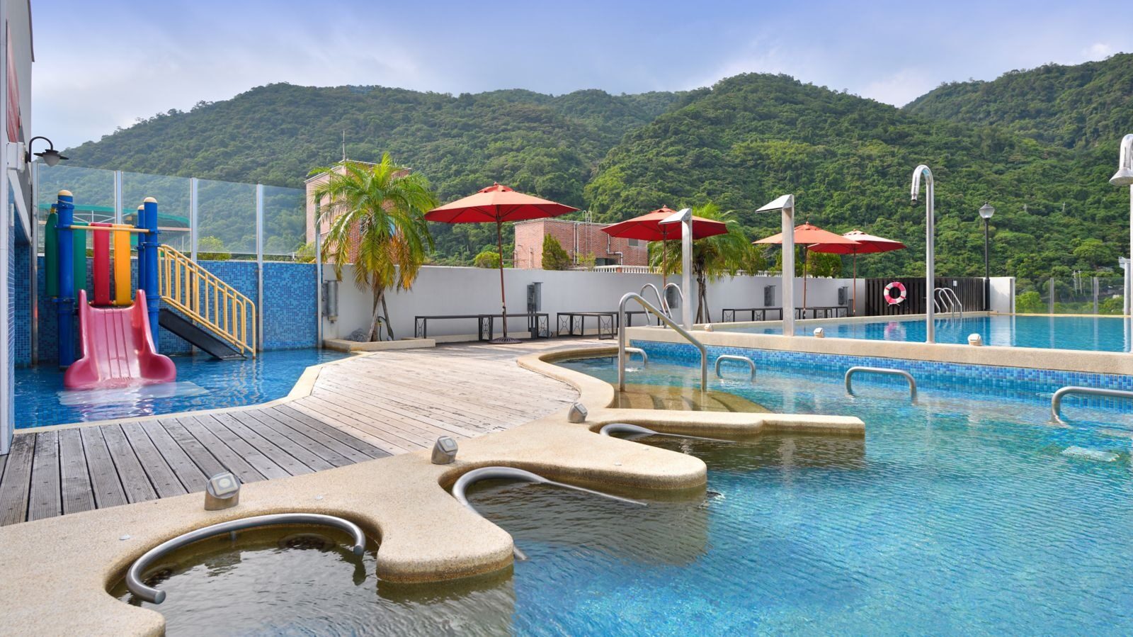 An outdoor pool beneath the mountains, with a pink slide and red sun umbrellas by the water