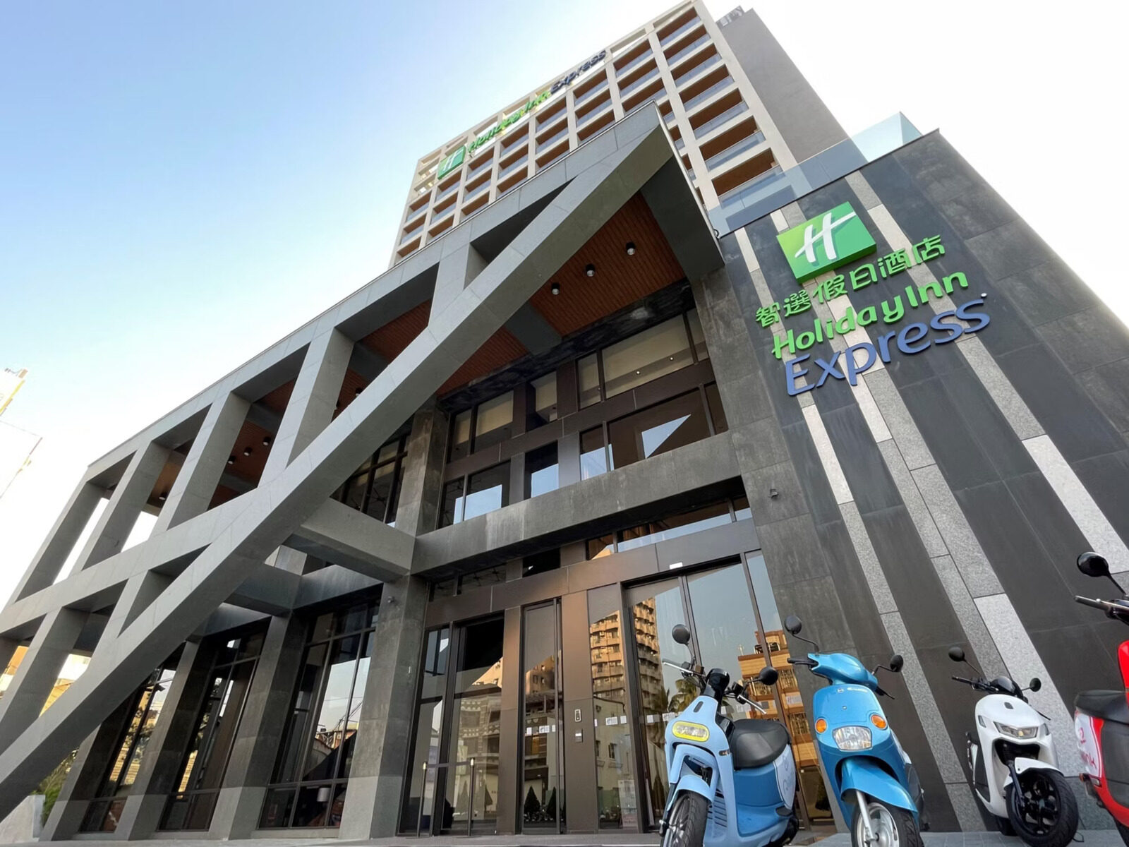Looking up at the hotel entrance from the street, a Holiday Inn Express sign on the gray grid facade and scooters parked out front