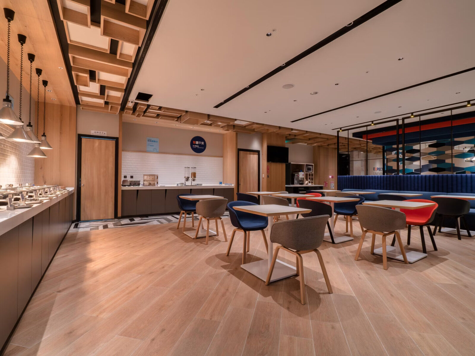 A breakfast area with wooden floors, a row of tables and chairs beside a long buffet counter and pendant lights
