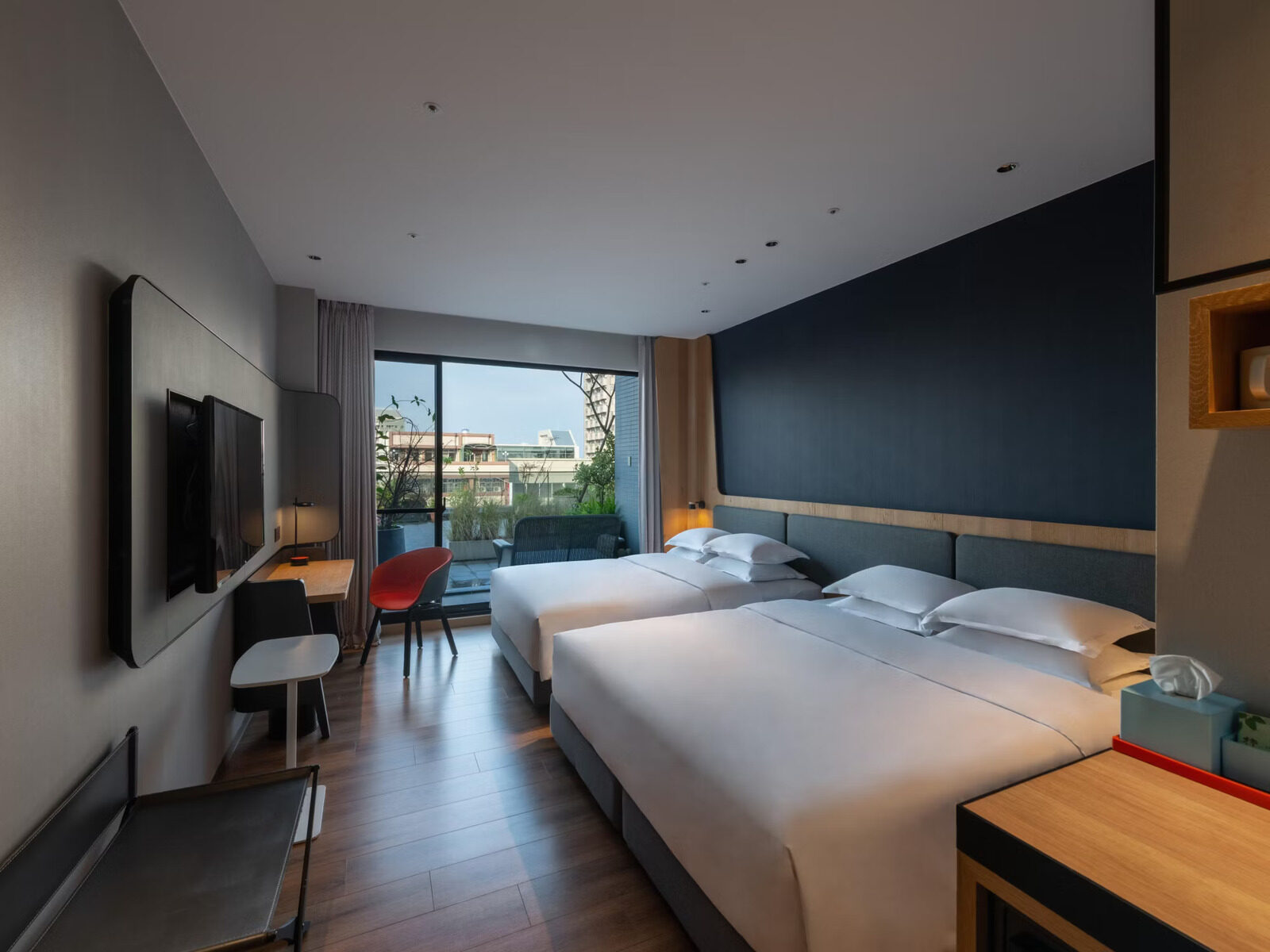A room with two large beds side by side, a balcony and green trees outside the window and a wall-mounted TV