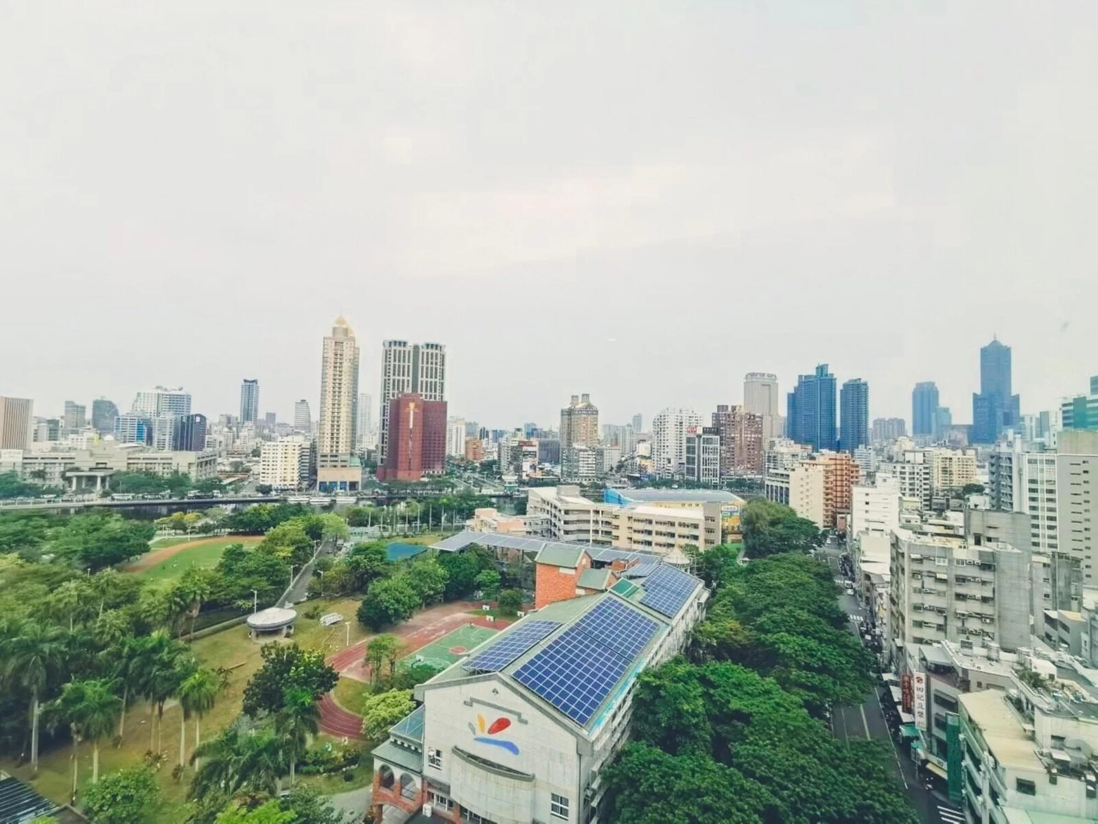 从高楼层望出去的高雄市区，前景是一片绿树与屋顶，远方是成排高楼