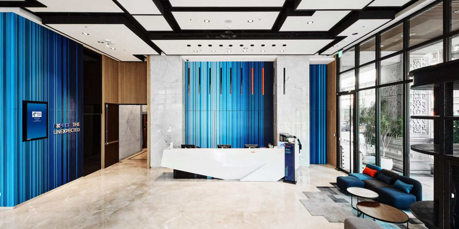 A white reception desk in the double-height lobby, backed by a blue striped wall, with sofas by the floor-to-ceiling windows on the right