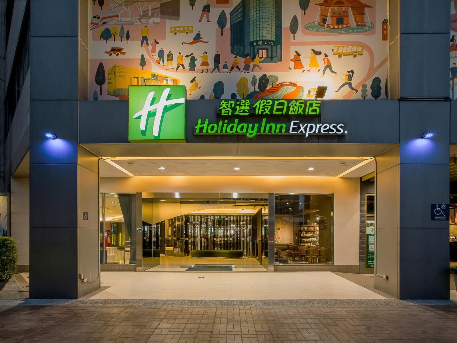 The hotel entrance lit up at night, with the green Holiday Inn Express sign and a colorful street-scene mural above the canopy