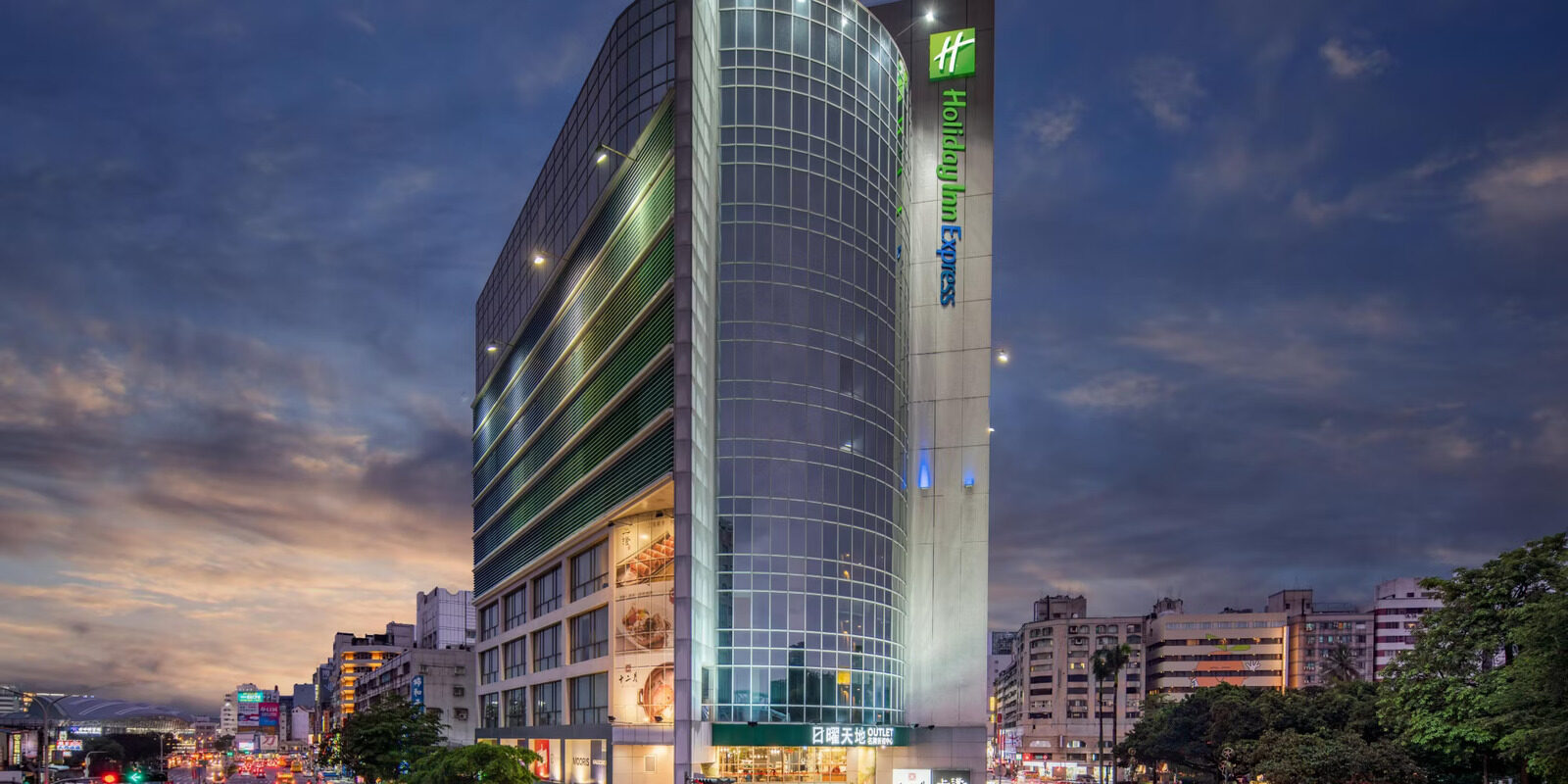 A glass-curtain-wall tower lit up at dusk, with a vertical Holiday Inn Express sign at the top and a city street in front