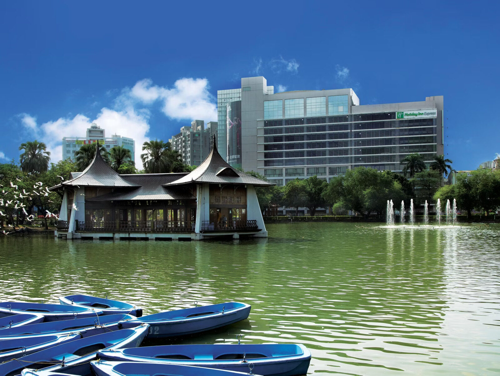 The Mid-Lake Pavilion in Taichung Park standing over the lake, with blue rowboats in front and the hotel sign atop a tower behind