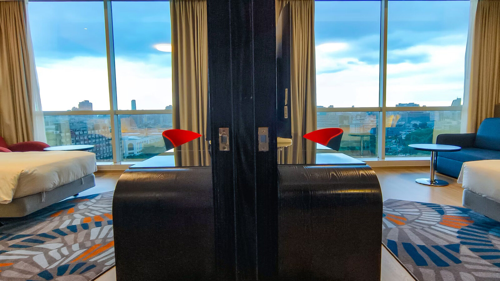 The connecting door between two rooms standing open, with a bed on each side and city views through the floor-to-ceiling windows