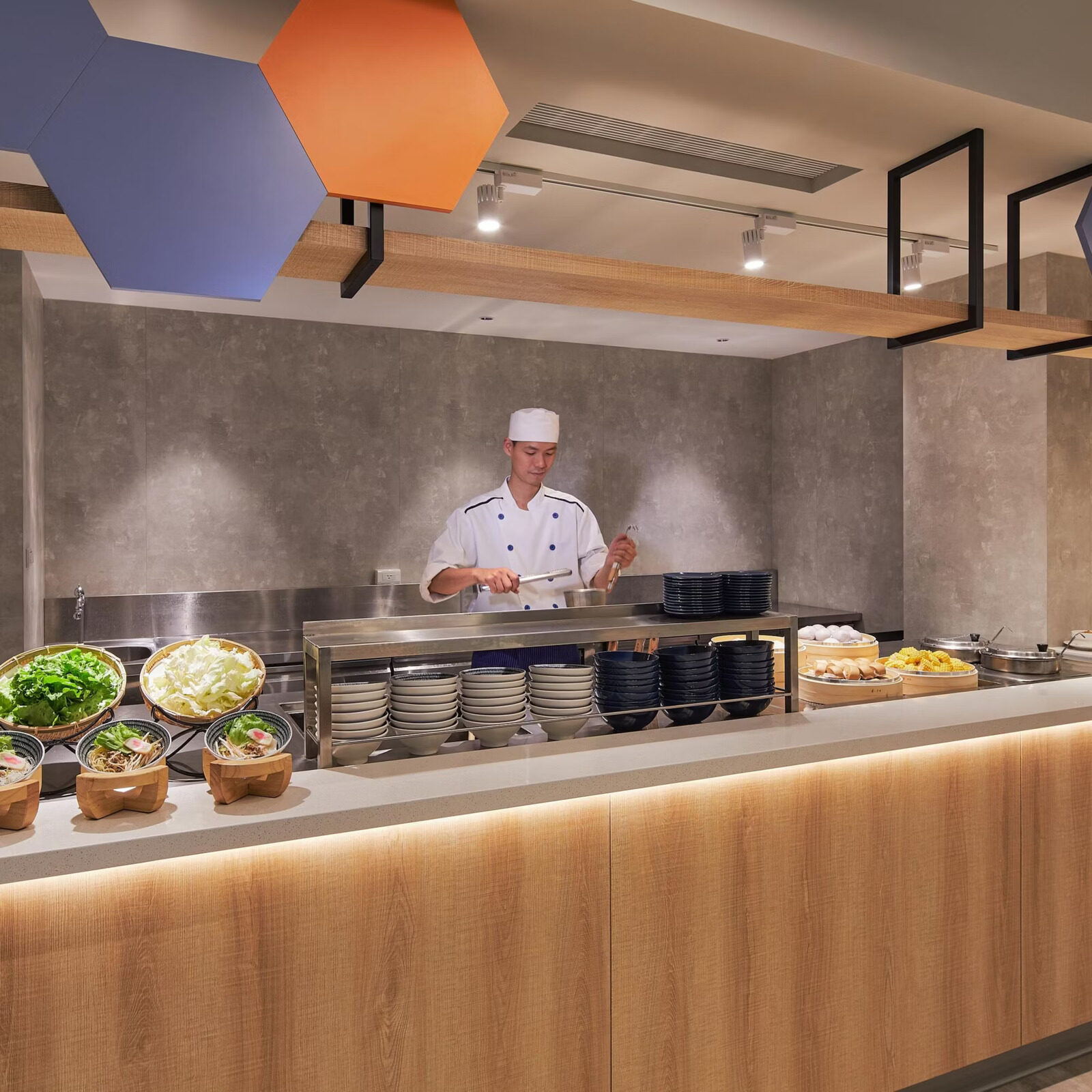 A chef in white standing behind the noodle soup station, with toppings, bowls and plates on the counter