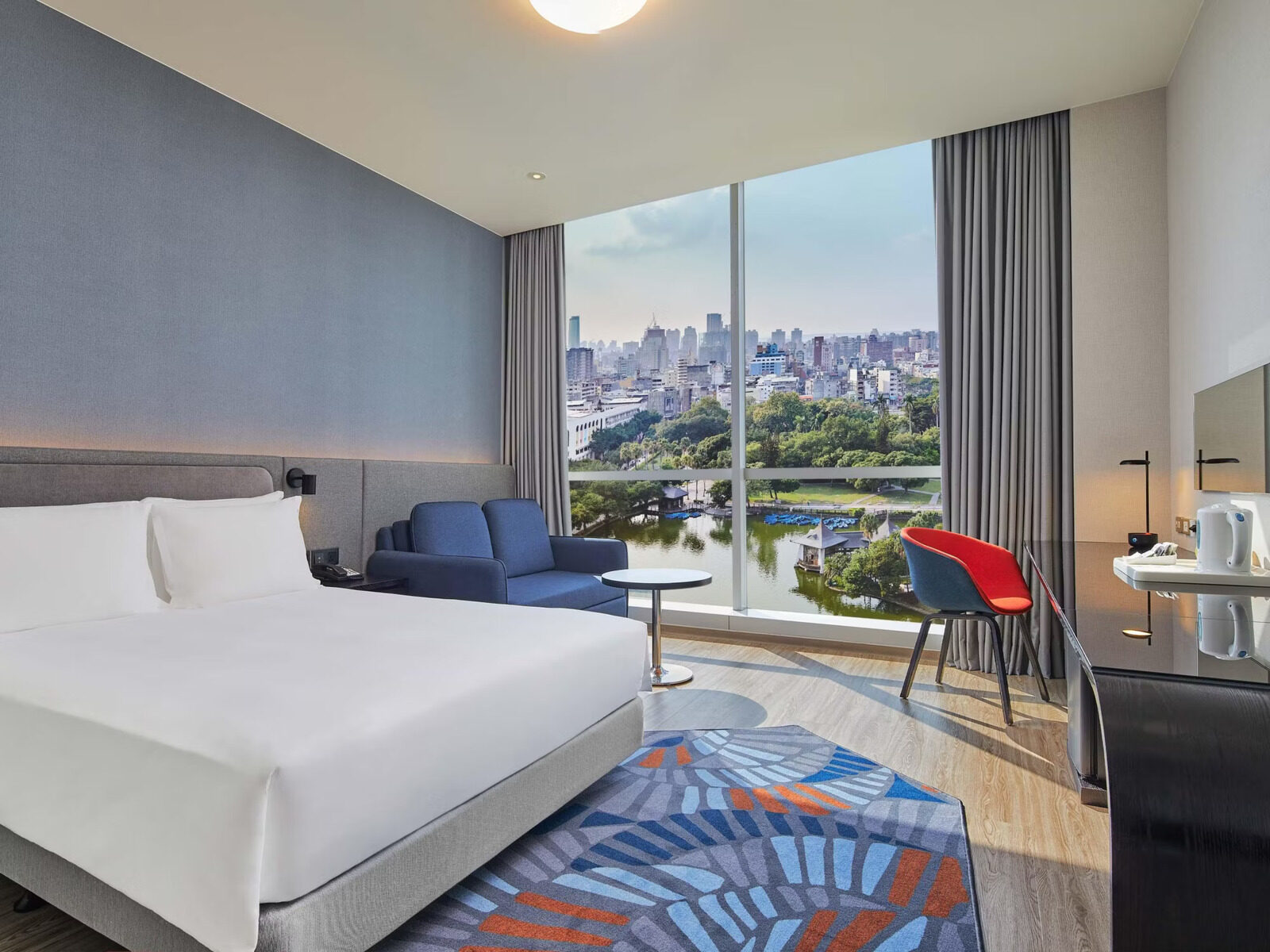 A king room with Taichung Park's trees and lake beyond the floor-to-ceiling window, and city high-rises in the distance