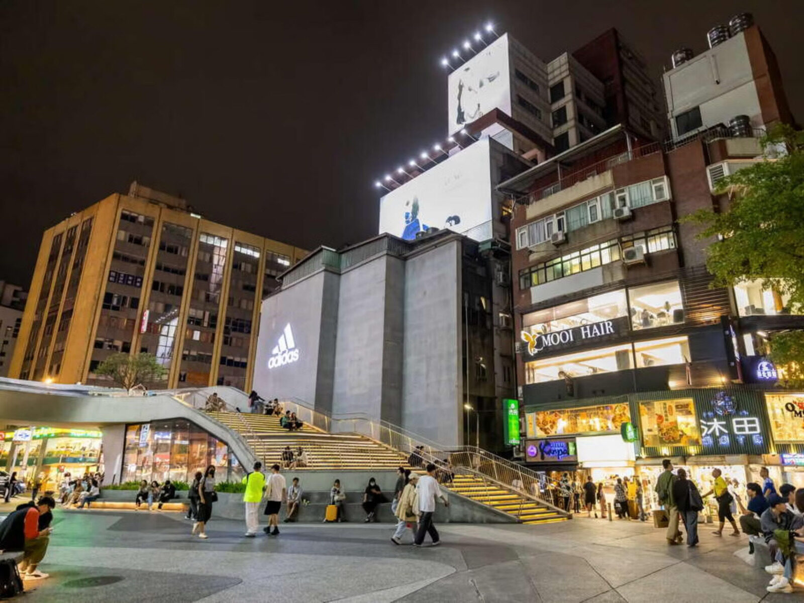 夜晚的街口，阶梯广场旁是亮灯的商店招牌与往来人潮