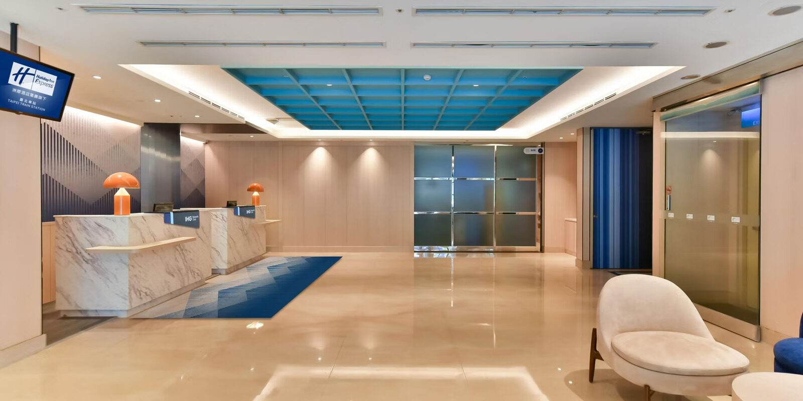 A bright hotel lobby, with a white stone reception desk in front of a blue-lit ceiling and glass doors