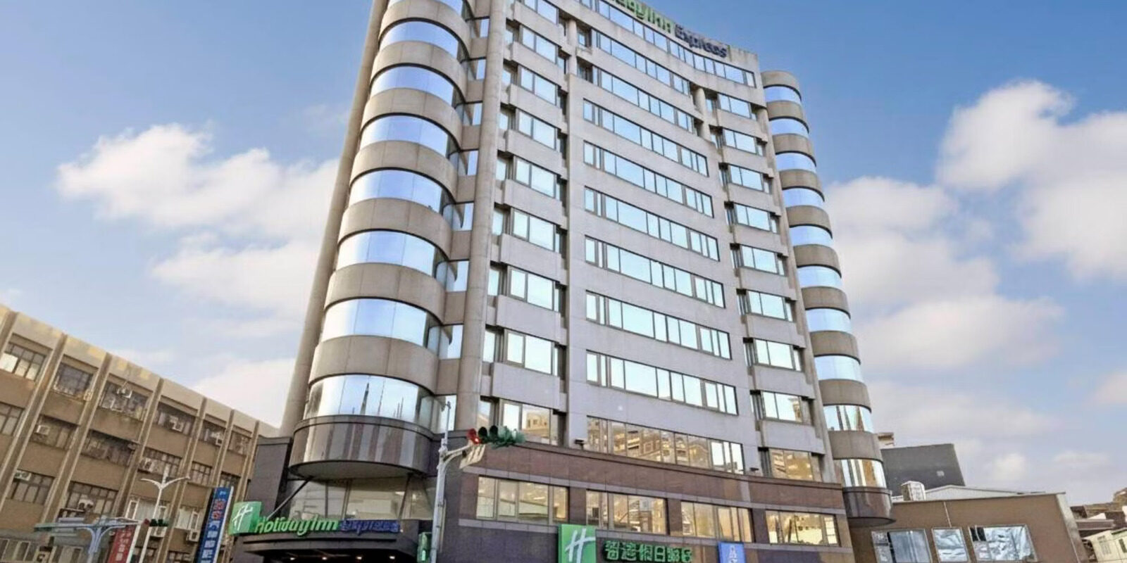 Looking up from the street at the hotel tower, with rows of windows on its curved facade against blue sky and white clouds