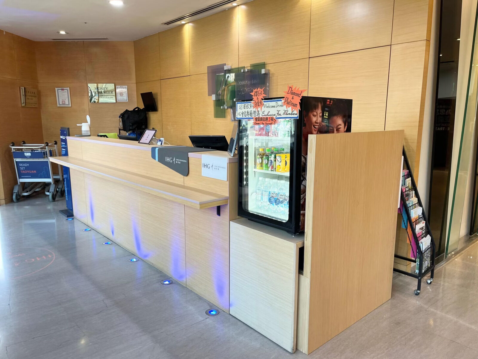 A light wood-grain reception counter next to a chilled drinks fridge and a luggage cart