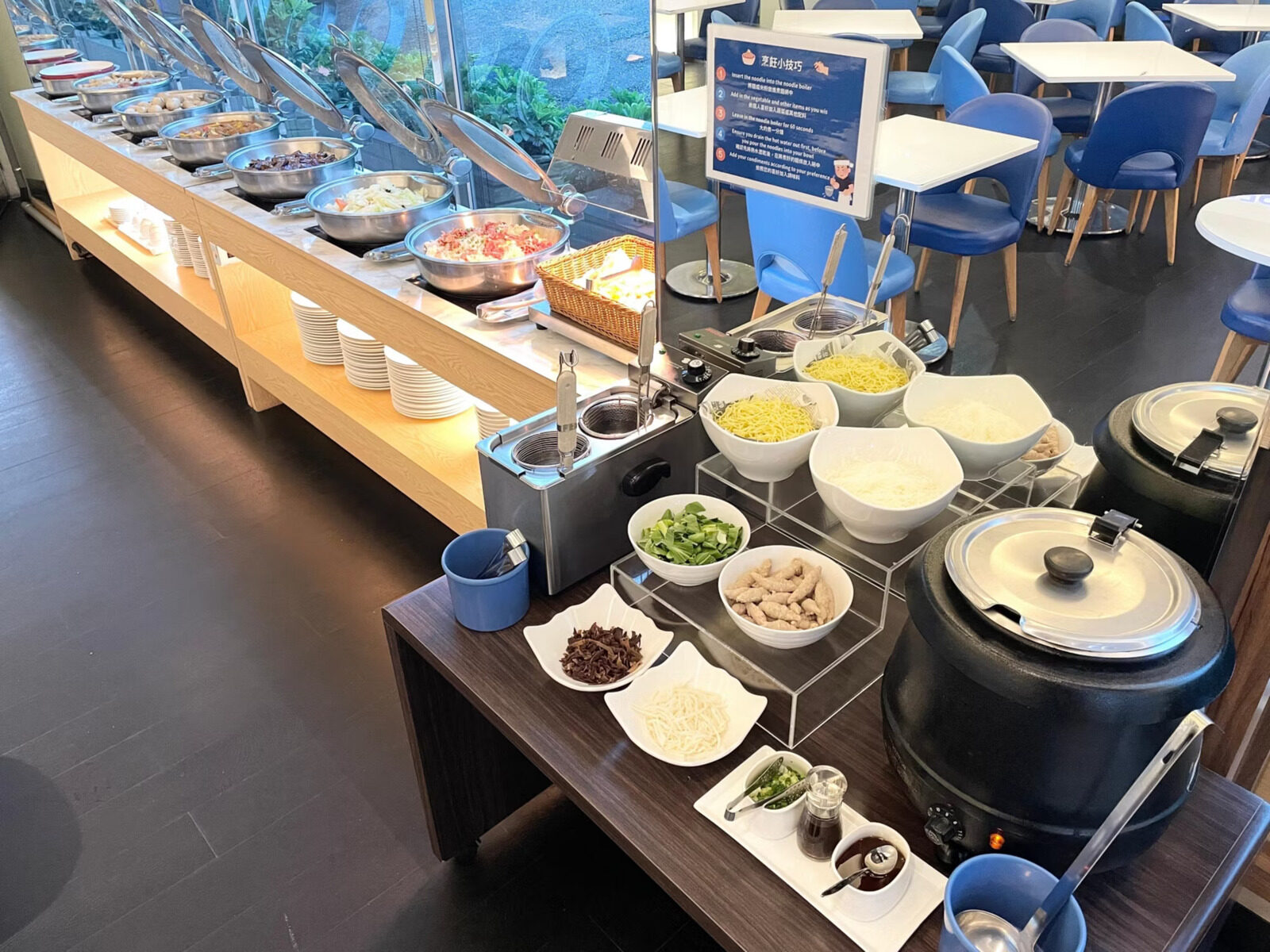 A make-your-own noodle station with noodles, greens and toppings next to a soup pot, with a row of chafing dishes and a dining area of blue chairs behind