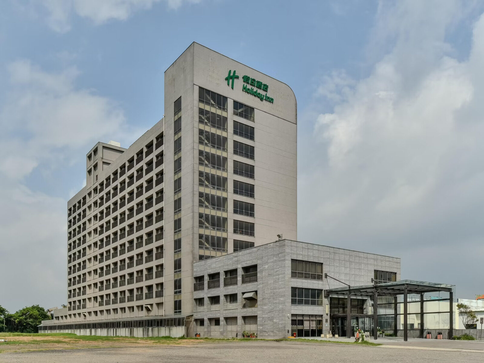 A gray hotel tower with a green Holiday Inn logo near the top, a glass-canopied entrance on the lower floors to the right and an open lot in front