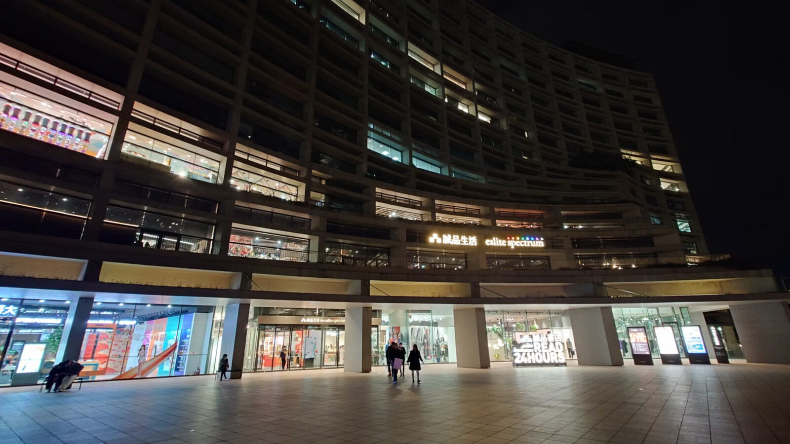 夜晚的诚品生活大楼，弧形立面亮着灯，一楼是整排店面，广场上有行人走过