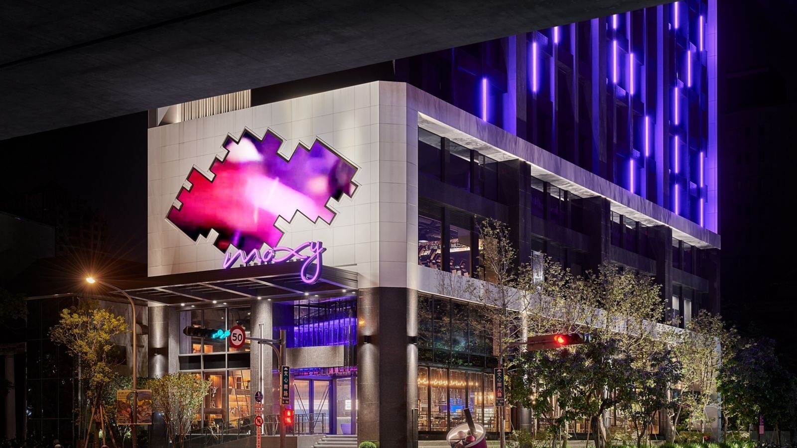 夜晚街角的酒店低楼层，外墙挂着桃红色 Moxy 图案灯箱，旁边是行道树