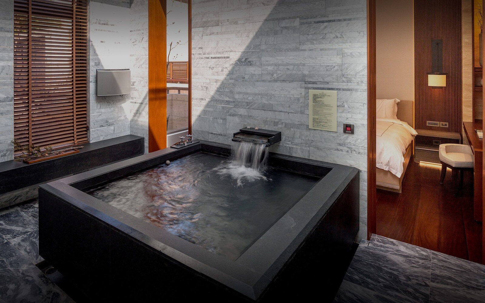 A square black stone hot spring bath and spout in a room, with the bedroom alongside